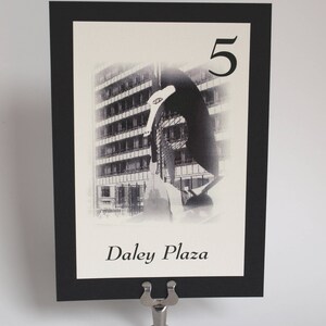 May include: Black and white photo of a building with a sculpture in front of it. The photo is on a black card with a white border. The card has the number "5" and the text "Daley Plaza" printed on it.