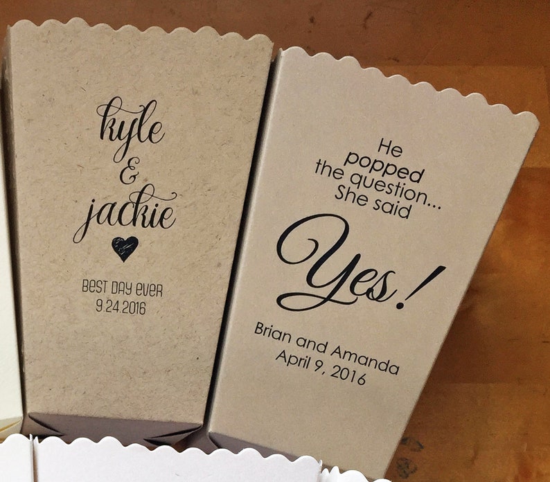 Pode incluir: Duas caixas de pipoca de papel castanho com texto. A primeira caixa diz "kyle & jackie" com um cora&ccedil;&atilde;o e "BEST DAY EVER 9.24.2016". A segunda caixa diz "He popped the question... She said Yes!" com "Brian and Amanda April 9, 2016" abaixo.