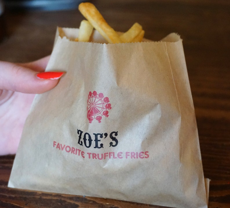 Op de afbeelding: Een bruine papieren zak met de tekst "ZOE'S FAVORITE TRUFFLE FRIES" erop gedrukt. Boven de tekst staat een rood reuzenradlogo. De zak is gevuld met friet.