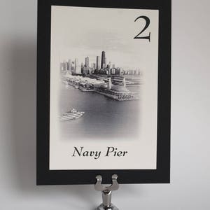 May include: Black and white photo of the Chicago skyline with the text "Navy Pier" and the number "2" on a white card with a black border. The card is in a silver table number holder.
