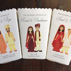 以下が含まれることがあります： インドの伝統的な結婚式衣装を着たカップルのイラストが描かれた白い箱が3つあります。箱には「The Wedding Ceremony of」という文字と、カップルの名前、結婚式の日付と場所が書かれています。