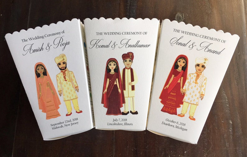 以下が含まれることがあります： インドの伝統的な結婚式衣装を着たカップルのイラストが描かれた白い箱が3つあります。箱には「The Wedding Ceremony of」という文字と、カップルの名前、結婚式の日付と場所が書かれています。