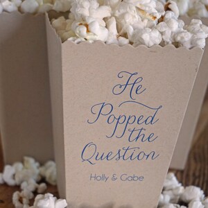 Pode incluir: Uma caixa de pipoca de papel castanho com o texto "He Popped the Question" em escrita azul e os nomes "Holly & Gabe" abaixo. A caixa est&aacute; cheia de pipocas brancas e rodeada por mais pipocas.