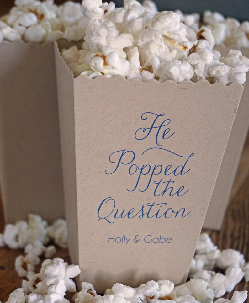 Pode incluir: Uma caixa de pipoca de papel castanho com o texto "He Popped the Question" em escrita azul e os nomes "Holly & Gabe" abaixo. A caixa est&aacute; cheia de pipocas brancas e rodeada por mais pipocas.