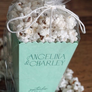 Pu&ograve; includere: Una scatola di popcorn verde chiaro con il testo "Angelina & Charley" e "Settembre 1925" stampato su di essa. La scatola &egrave; piena di popcorn e avvolta in plastica trasparente.