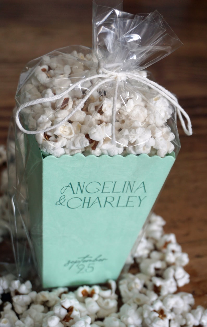Pu&ograve; includere: Una scatola di popcorn verde chiaro con il testo "Angelina & Charley" e "Settembre 1925" stampato su di essa. La scatola &egrave; piena di popcorn e avvolta in plastica trasparente.