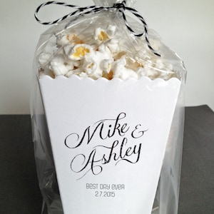 May include: A white popcorn container filled with popcorn, enclosed in a clear plastic bag tied with black and white twine. The container features the black printed text Mike & Ashley, followed by the text BEST DAY EVER and the date 2.7.2015.