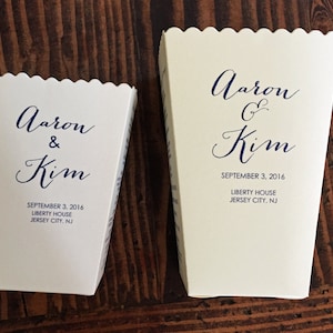 Peut inclure: Deux boîtes à popcorn blanches avec des bords festonnés. Les boîtes sont décorées d'une calligraphie bleue qui dit "Aaron & Kim" et "Aaron G Kim". Les boîtes comprennent également la date "3 septembre 2016", le lieu "Liberty House" et la ville "Jersey City, NJ".