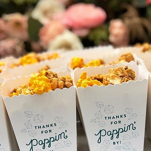 May include: White paper popcorn containers with scalloped edges filled with yellow and brown popcorn. The front of each container features the text "THANKS FOR Poppin' BY!" in dark blue, surrounded by small geometric diamond-shaped line drawings.