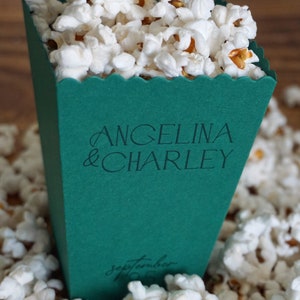 Pu&ograve; includere: Una scatola di popcorn verde con un bordo a zig zag. La scatola &egrave; decorata con i nomi "Angelina & Charley" e la data "14 settembre 2023".