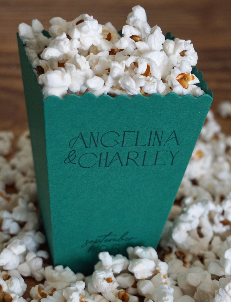 Pu&ograve; includere: Una scatola di popcorn verde con un bordo a zig zag. La scatola &egrave; decorata con i nomi "Angelina & Charley" e la data "14 settembre 2023".