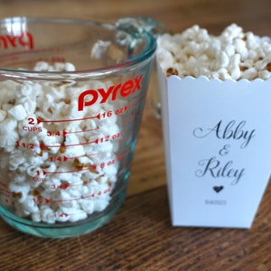 May include: A clear glass Pyrex measuring cup filled with white popcorn next to a white paper popcorn container. The container features the text Abby & Riley in a black script font with a small heart icon and the date 8/4/2023 printed below.