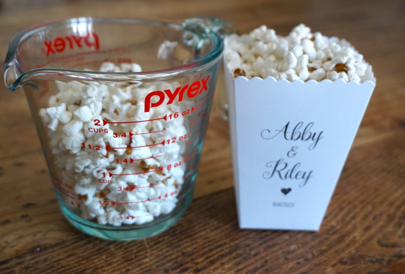 May include: A clear glass Pyrex measuring cup filled with white popcorn next to a white paper popcorn container. The container features the text Abby & Riley in a black script font with a small heart icon and the date 8/4/2023 printed below.