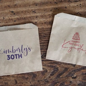 Op de afbeelding: Twee bruine papieren zakken met aangepaste bedrukte tekst. De eerste zak zegt "Kimberly's 30th" in paarse schrijfletters. De tweede zak zegt "Happy Fry-Day 2 Kyndall" in rode schrijfletters met een rood taartpictogram.