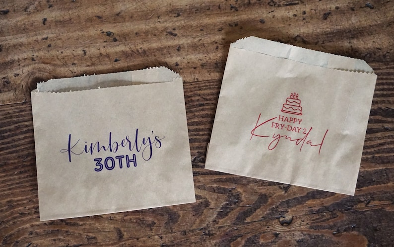 Op de afbeelding: Twee bruine papieren zakken met aangepaste bedrukte tekst. De eerste zak zegt "Kimberly's 30th" in paarse schrijfletters. De tweede zak zegt "Happy Fry-Day 2 Kyndall" in rode schrijfletters met een rood taartpictogram.
