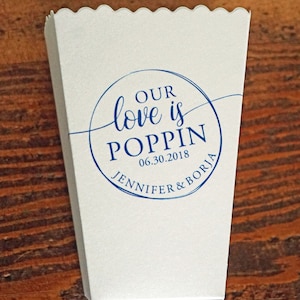 May include: A white paper popcorn container with a scalloped top edge. The front features a blue circular graphic with the text "OUR love is POPPIN", the date "06.30.2018", and the names "JENNIFER & BORJA" printed in a mix of script and serif fonts.