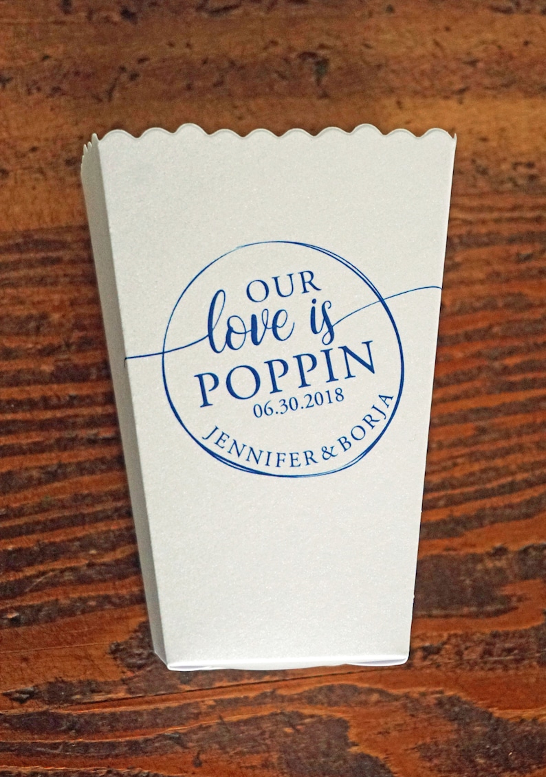 May include: A white paper popcorn container with a scalloped top edge. The front features a blue circular graphic with the text "OUR love is POPPIN", the date "06.30.2018", and the names "JENNIFER & BORJA" printed in a mix of script and serif fonts.