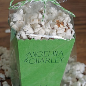 Pu&ograve; includere: Una scatola di popcorn verde con un fiocco di corda bianca e un sacchetto di plastica trasparente. La scatola &egrave; decorata con il testo "Angelina & Charley" e la data "September 25".