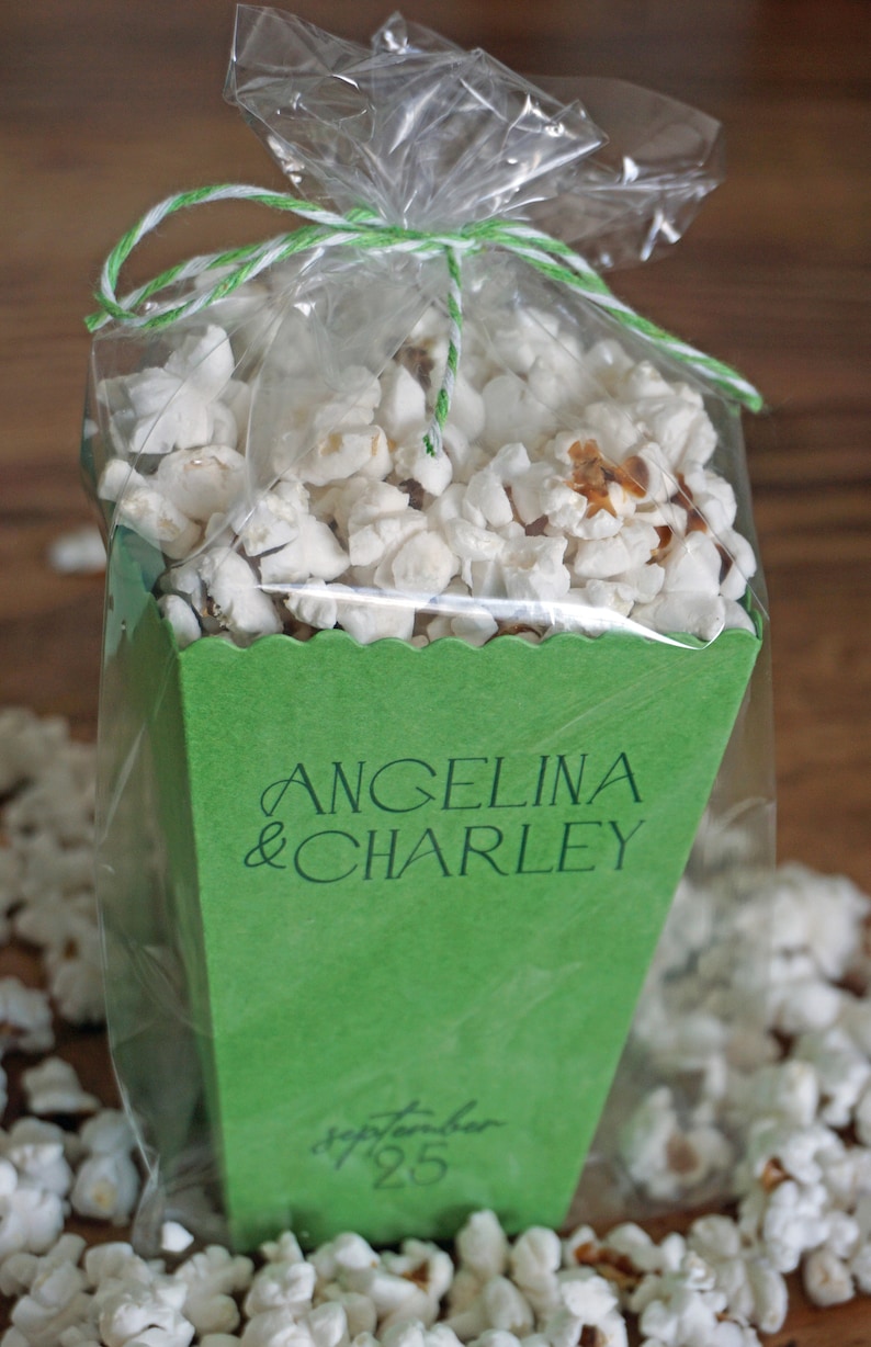 Pu&ograve; includere: Una scatola di popcorn verde con un fiocco di corda bianca e un sacchetto di plastica trasparente. La scatola &egrave; decorata con il testo "Angelina & Charley" e la data "September 25".
