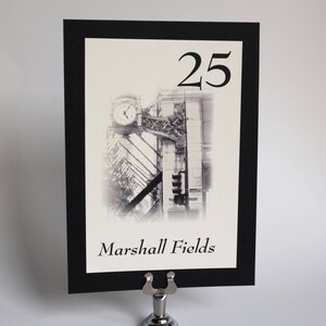May include: Black and white photo of a building with a clock tower. The number 25 is printed above the photo. The text "Marshall Fields" is printed below the photo. The photo is framed with a black border and is displayed on a silver stand.