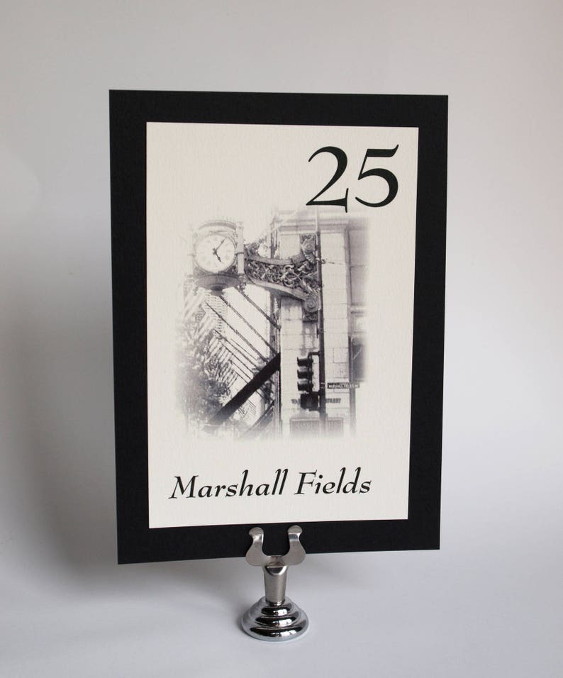 May include: Black and white photo of a building with a clock tower. The number 25 is printed above the photo. The text "Marshall Fields" is printed below the photo. The photo is framed with a black border and is displayed on a silver stand.