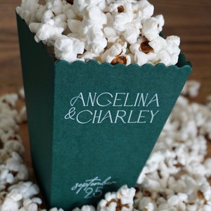 Pu&ograve; includere: Una scatola di popcorn verde con testo bianco che dice "Angelina & Charley" e "Settembre 25". La scatola &egrave; piena di popcorn bianchi e circondata da altro popcorn.
