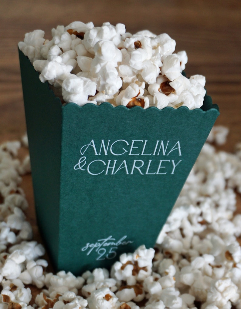 Pu&ograve; includere: Una scatola di popcorn verde con testo bianco che dice "Angelina & Charley" e "Settembre 25". La scatola &egrave; piena di popcorn bianchi e circondata da altro popcorn.