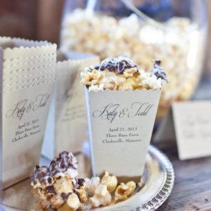 May include: Off-white paper popcorn containers with scalloped edges, printed with the text Katy & Todd, April 27, 2013, Three Barn Farm, Clarksville, Minnesota. The containers are filled with popcorn drizzled with dark chocolate on a silver tray.