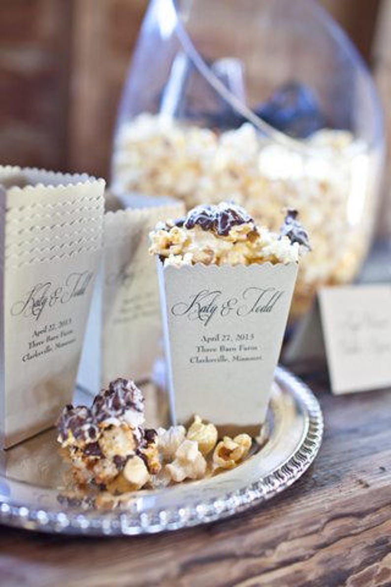 May include: Off-white paper popcorn containers with scalloped edges, printed with the text Katy & Todd, April 27, 2013, Three Barn Farm, Clarksville, Minnesota. The containers are filled with popcorn drizzled with dark chocolate on a silver tray.