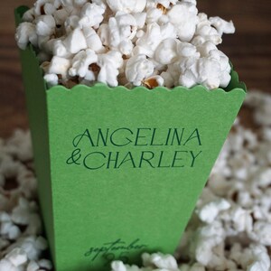 Pu&ograve; includere: Una scatola di popcorn verde con il testo "Angelina & Charley" e "Settembre 19" stampato su di essa. La scatola &egrave; piena di popcorn bianchi e circondata da altro popcorn.