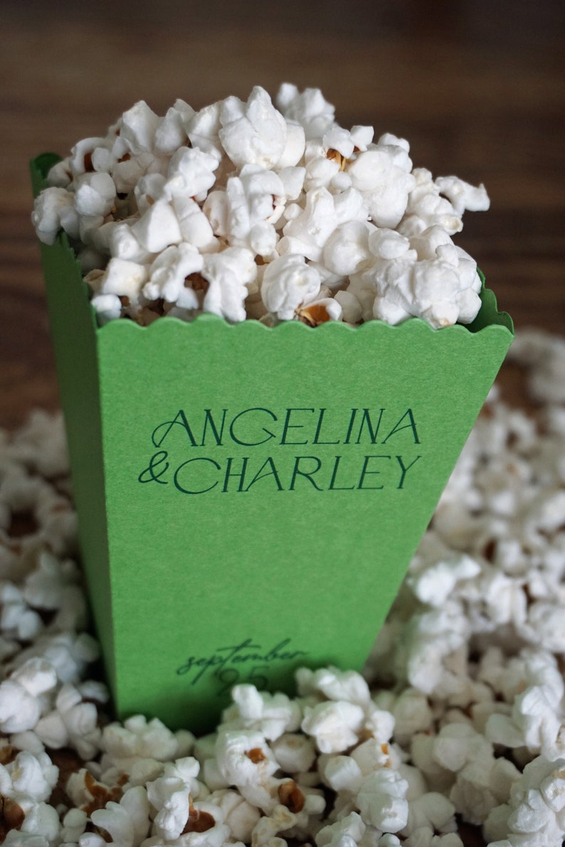 Pu&ograve; includere: Una scatola di popcorn verde con il testo "Angelina & Charley" e "Settembre 19" stampato su di essa. La scatola &egrave; piena di popcorn bianchi e circondata da altro popcorn.