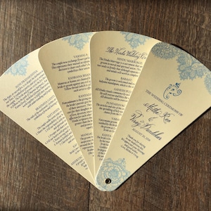 May include: A cream-colored wedding program fan with blue floral designs and text. The fan is open, displaying several panels with event details and the names of the couple, Mithu Rao and Virag Dandekar, with the date August 20, 2018.