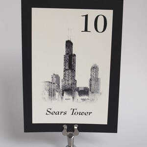 May include: Black and white illustration of the Sears Tower, also known as Willis Tower, in Chicago. The illustration is on a white card with a black border and the number 10 printed in black at the top. The card is on a silver table number holder.