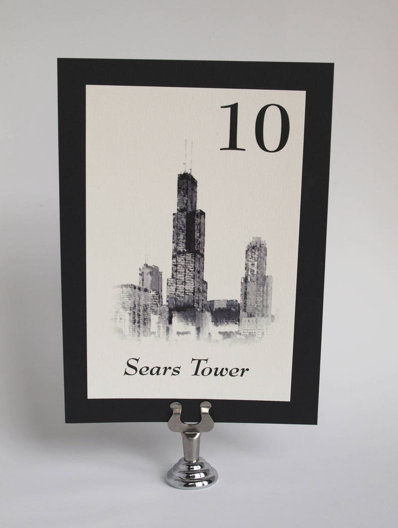 May include: Black and white illustration of the Sears Tower, also known as Willis Tower, in Chicago. The illustration is on a white card with a black border and the number 10 printed in black at the top. The card is on a silver table number holder.