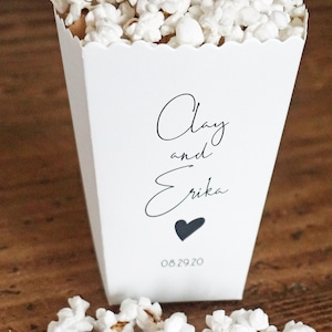 May include: A white paper popcorn container with a scalloped top edge, filled with white popcorn. The front features black text reading Clay and Erika, a small black heart icon, and the date 08.29.20 printed in a simple, clean font style.