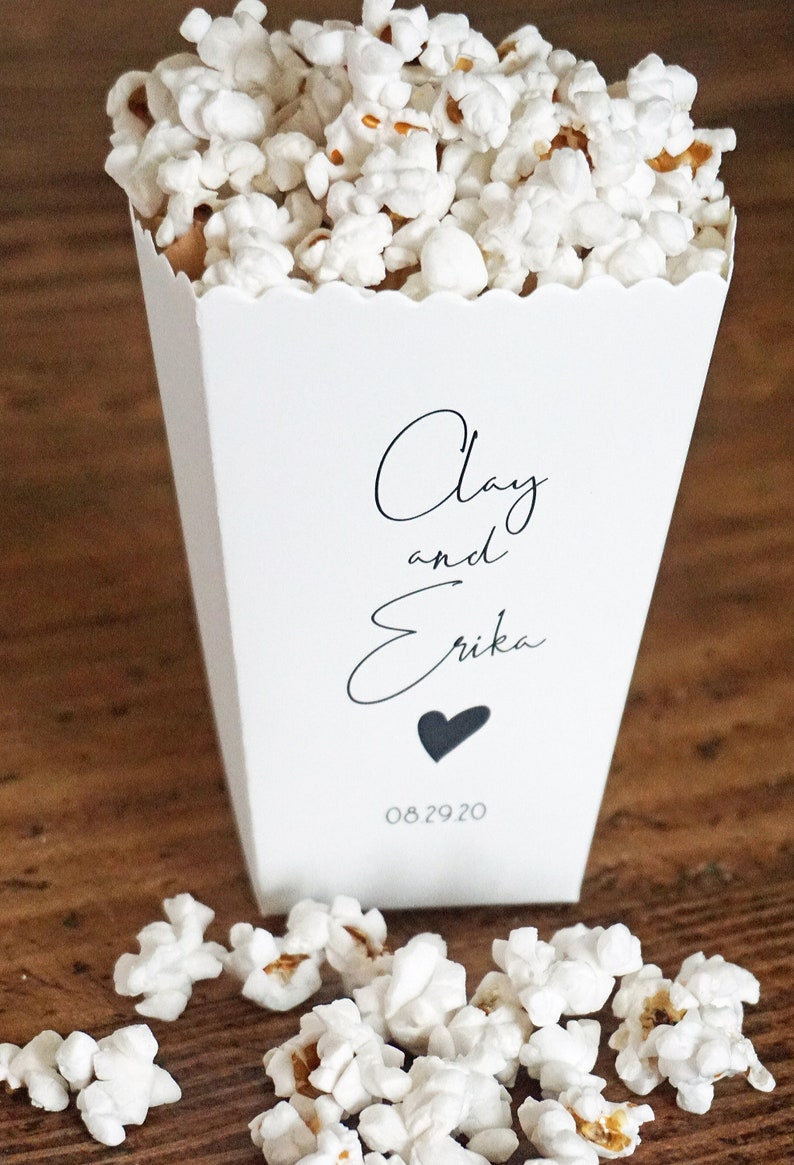 May include: A white paper popcorn container with a scalloped top edge, filled with white popcorn. The front features black text reading Clay and Erika, a small black heart icon, and the date 08.29.20 printed in a simple, clean font style.