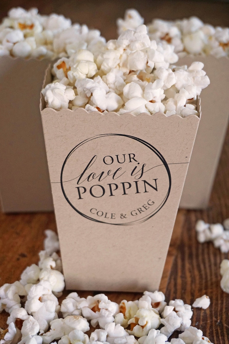 Pode incluir: Uma caixa de pipoca de papel castanho com um c&iacute;rculo preto e o texto "OUR love is POPPIN COLE & GREG". A caixa est&aacute; cheia de pipocas brancas e h&aacute; pipocas espalhadas sobre uma superf&iacute;cie de madeira.