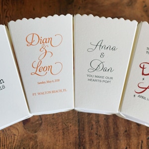 May include: Four off-white paper popcorn boxes with scalloped top edges, each featuring unique custom printed text and dates. The designs include names like Phung & Robin, Dian & Leon, Anna & Dan, and Destiny & Alex, along with various event dates and phrases.