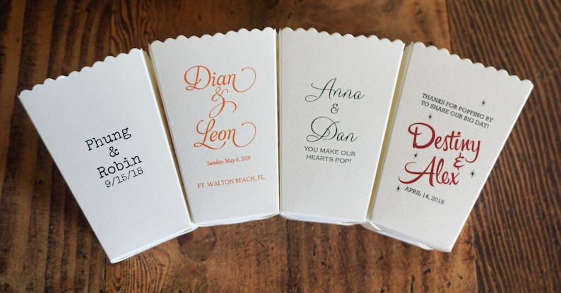 May include: Four off-white paper popcorn boxes with scalloped top edges, each featuring unique custom printed text and dates. The designs include names like Phung & Robin, Dian & Leon, Anna & Dan, and Destiny & Alex, along with various event dates and phrases.