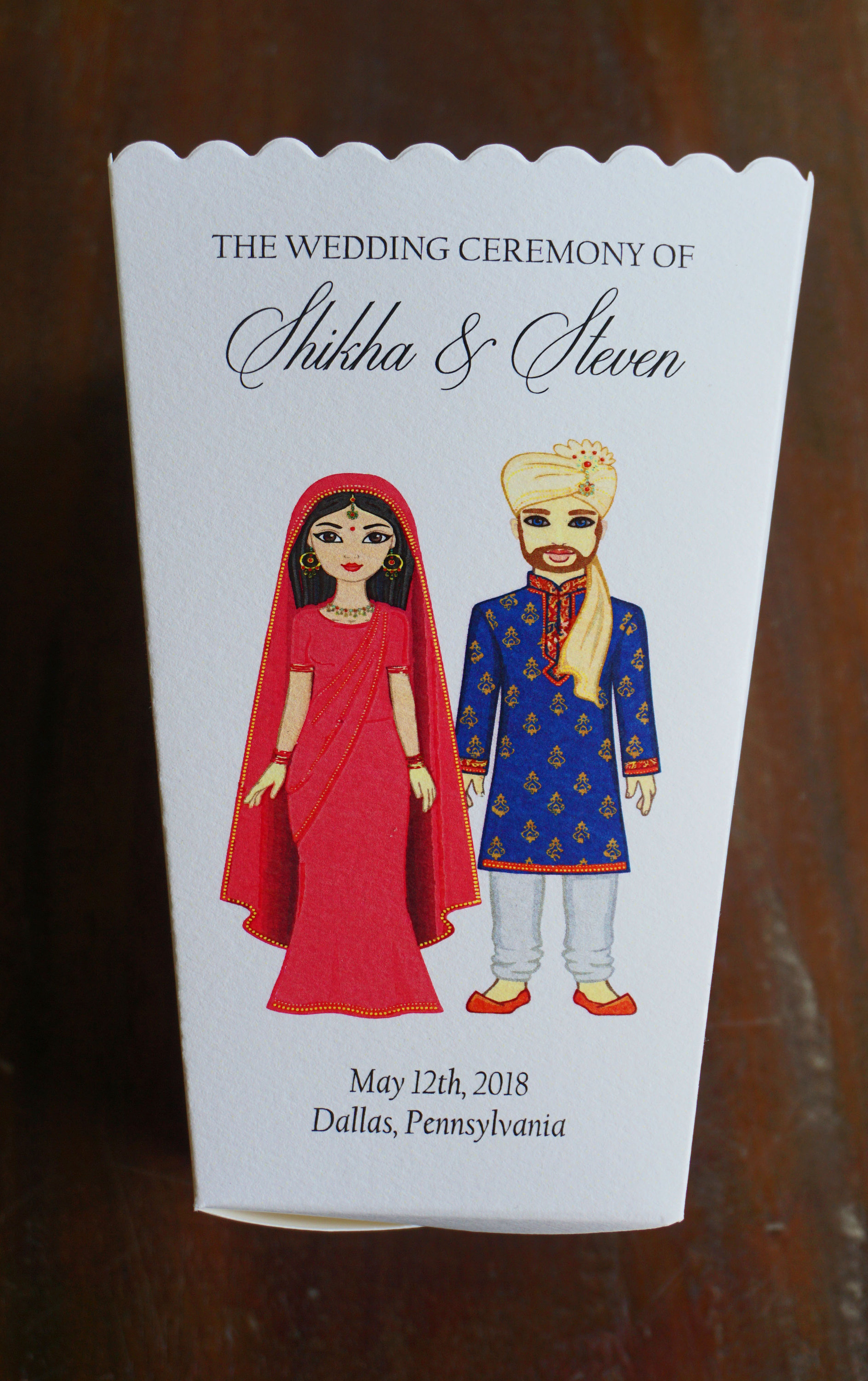 以下が含まれることがあります： シッハとスティーブンの結婚式を祝う招待状。インドの伝統的な衣装を着た新郎新婦の漫画イラストが描かれている。テキストには「The Wedding Ceremony of Shikha & Steven」と「May 12th, 2018 Dallas, Pennsylvania」と記載されている。