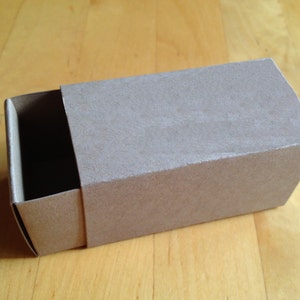 May include: A small, rectangular cardboard box with a sliding lid. The box is a light brown color and has a slightly textured surface.