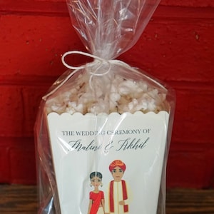 Puede incluir: Una caja de regalo blanca con borde festoneado, llena de palomitas de ma&iacute;z blancas. La caja tiene un anuncio de boda para Malini & Akhil, con una ilustraci&oacute;n de dibujos animados de la pareja con atuendo tradicional indio. El anuncio incluye la fecha "21 de enero de 2018" y la ubicaci&oacute;n "Rajkot, India".