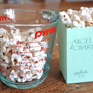 Pu&ograve; includere: Un misurino di vetro trasparente pieno di popcorn, con segni rossi per once e tazze. Accanto ad esso c'&egrave; una scatola di popcorn blu chiaro con il testo "ANGELINA & CHARLEY" e "Settembre 25".