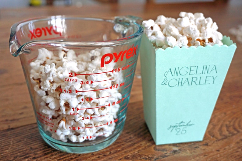 Pu&ograve; includere: Un misurino di vetro trasparente pieno di popcorn, con segni rossi per once e tazze. Accanto ad esso c'&egrave; una scatola di popcorn blu chiaro con il testo "ANGELINA & CHARLEY" e "Settembre 25".