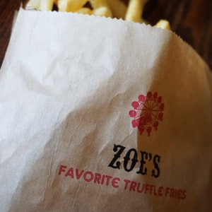 Op de afbeelding: Een bruine papieren zak met de tekst "ZOE'S FAVORITE TRUFFLE FRIES" erop gedrukt. Boven de tekst staat een rood en wit logo van een reuzenrad. De zak is gevuld met friet.