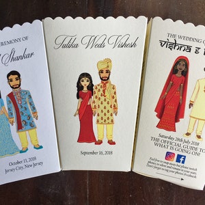 以下が含まれることがあります： インドの伝統的な衣装を着たカップルが描かれた、結婚式のお祝いの招待状が3枚。招待状は、波打つ縁取りのある白い厚紙に印刷されています。招待状には、カップルの名前、式典の日時、場所が記載されています。