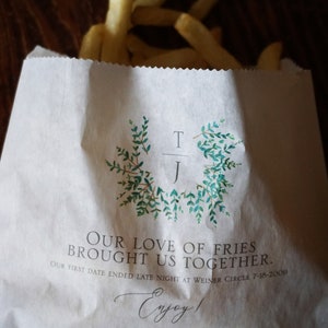 Op de afbeelding: Een witte papieren zak met een groene krans van bladeren en de initialen "T/J" erop gedrukt. De tekst "Our love of fries brought us together. Our first date ended late night at Weiner Circle 7-18-2009" staat onder de krans gedrukt. De tekst "Enjoy!" staat onder de datum gedrukt.