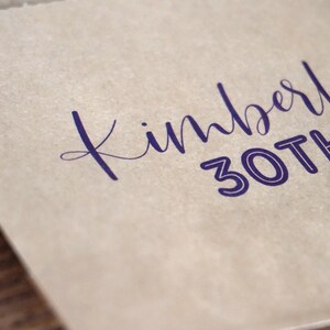 Op de afbeelding: Een bruine papieren zak met de tekst "Kimberl 30th" in paars gedrukt.