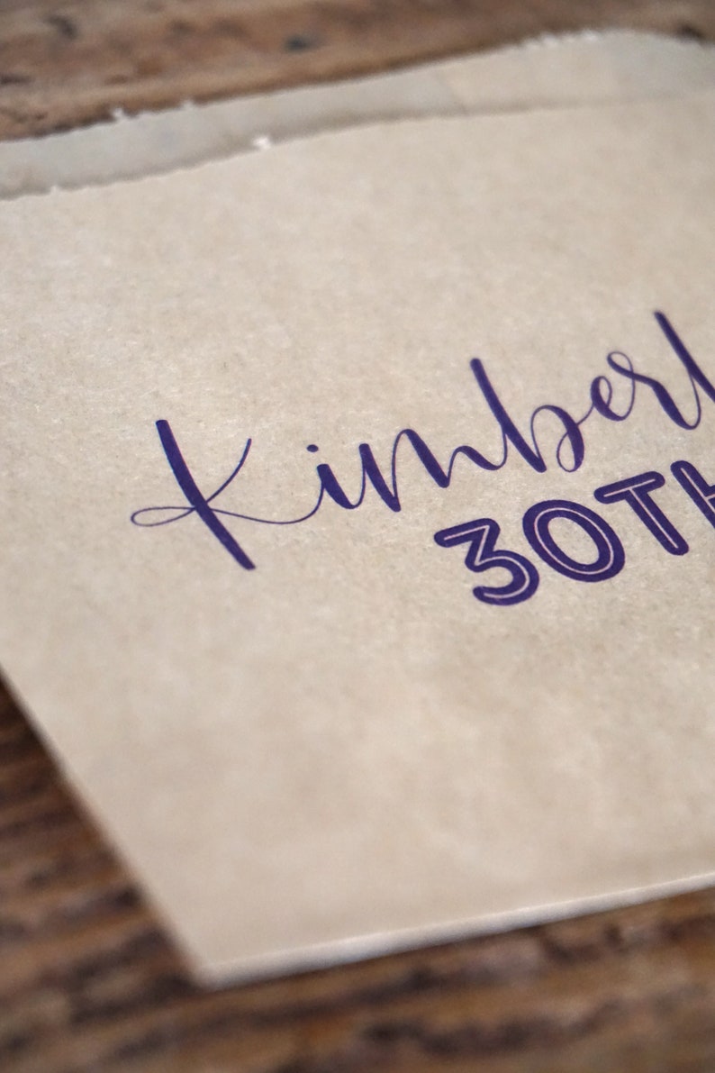 Op de afbeelding: Een bruine papieren zak met de tekst "Kimberl 30th" in paars gedrukt.