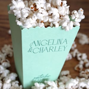 Pu&ograve; includere: Una scatola di popcorn verde chiaro con il testo "Angelina & Charley" stampato sul davanti. La scatola &egrave; piena di popcorn bianchi e circondata da altro popcorn.